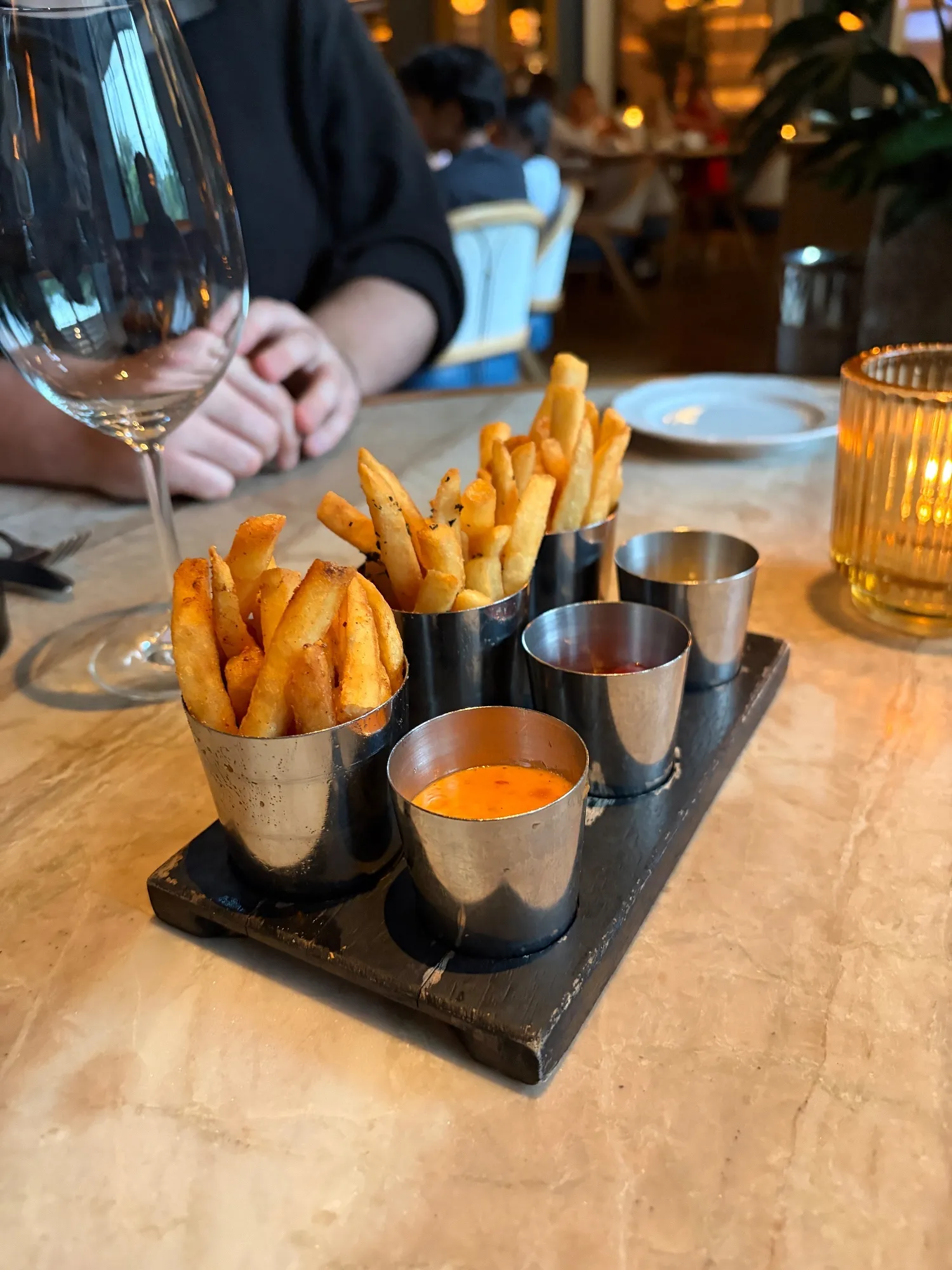 Several metal cups of French fries and dipping sauces sit on a wooden tray on a dining table.