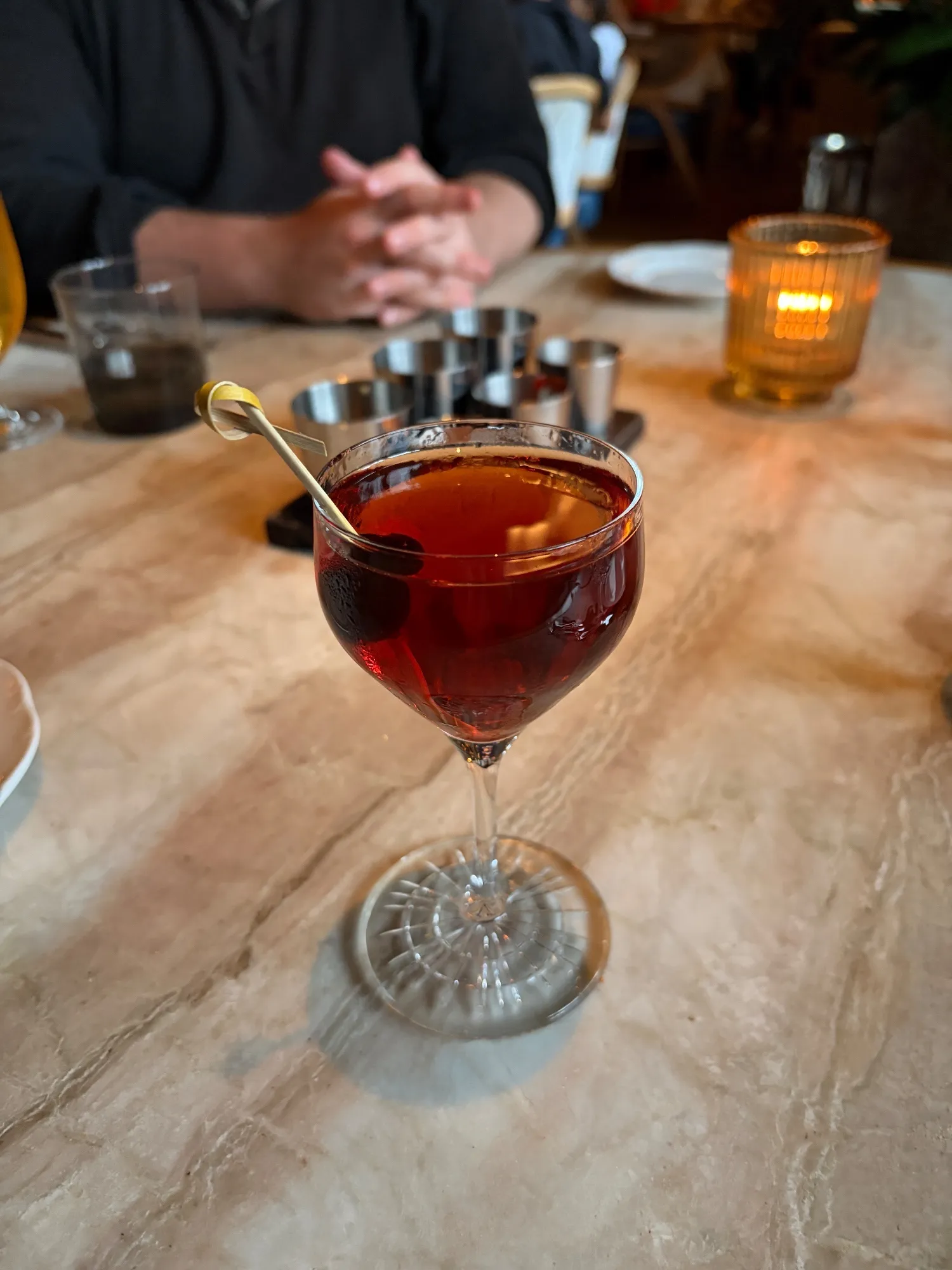 A red-brown cocktail with a skewered cherry sits in a stemmed glass on a marble table.