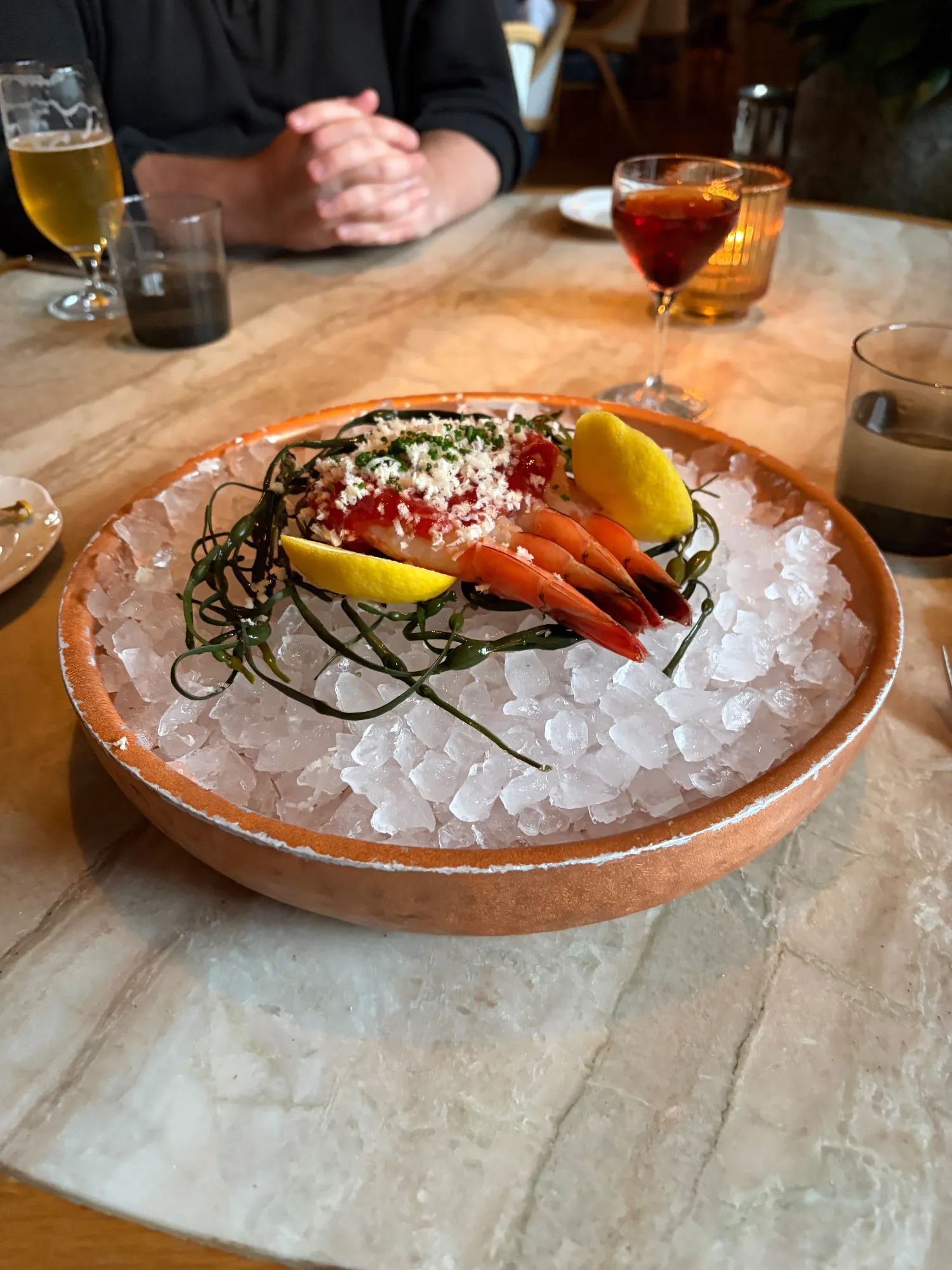 Four shrimp topped with red sauce and white shavings sit on crushed ice with lemon wedges and strands of seaweed.