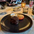 A round filet sits on a black plate beside a ramekin and a halved tomato topped with crumbs.