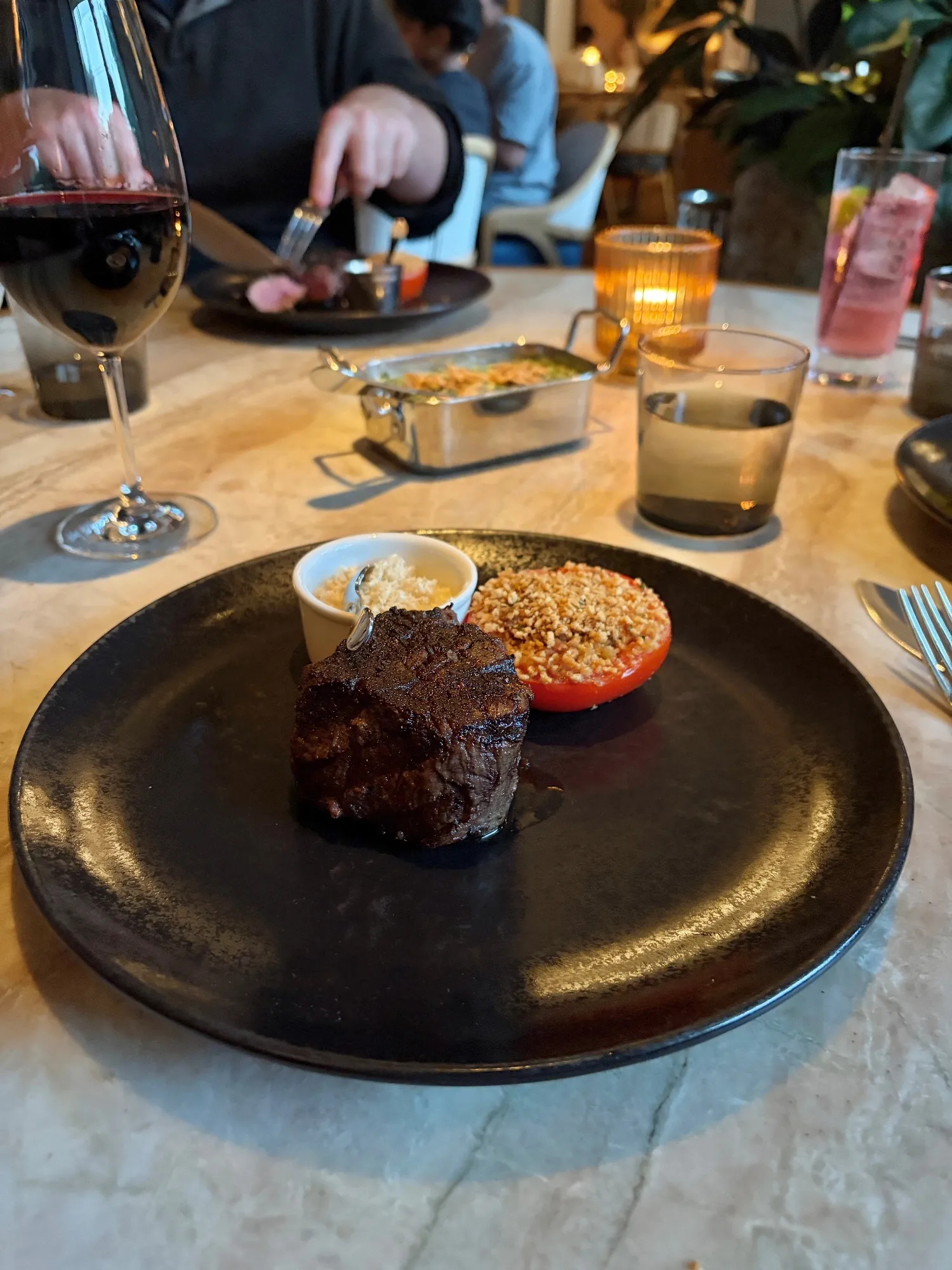 A round filet sits on a black plate beside a ramekin and a halved tomato topped with crumbs.