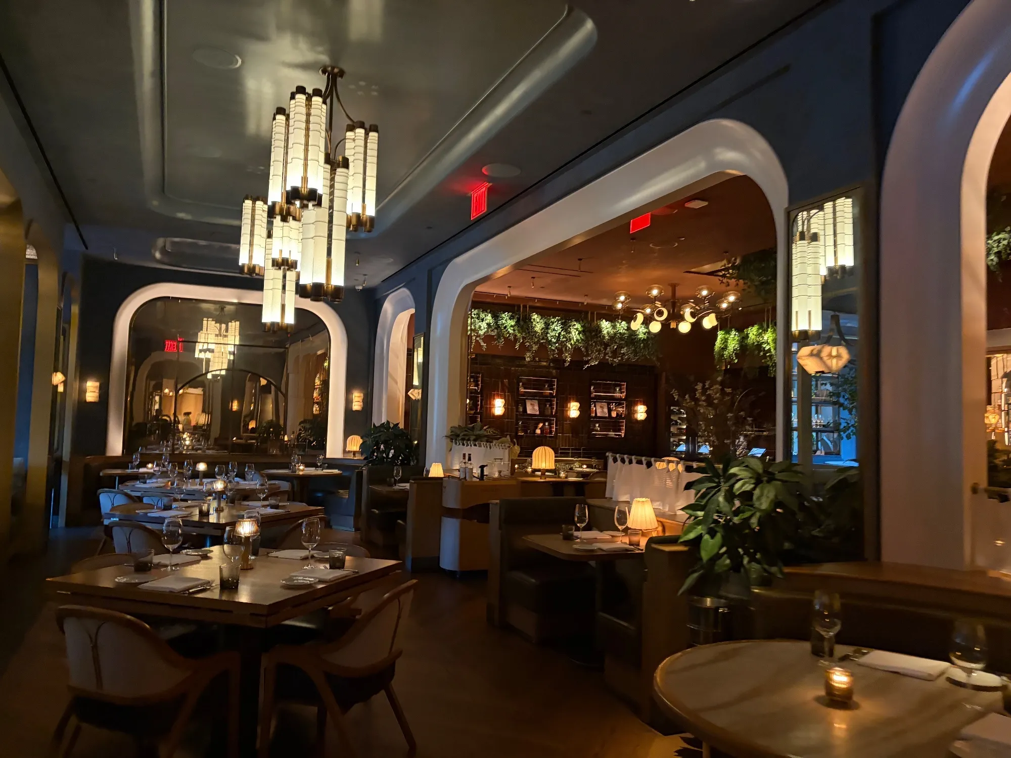 A dining room contains set tables, booths, arched openings, hanging lights and plants.