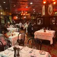 Dining room with wood paneling, framed paintings, patterned red carpet and warm overhead lighting, tables set with white cloths.