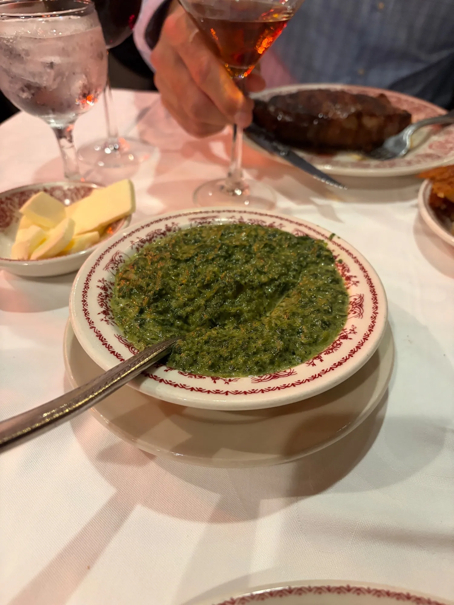 A bowl of creamed spinach with a spoon, a dish of butter behind it and a steak on a plate in the background.