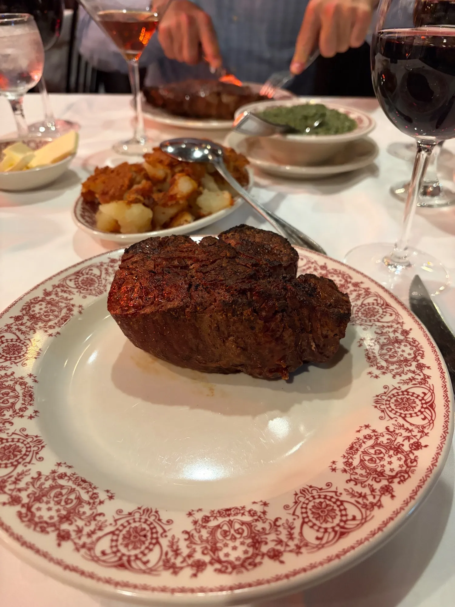 A seared filet on a red-patterned plate, with a side of hash browns and wine glasses behind.