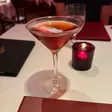 A brown cocktail served up in a stemmed glass on a white tablecloth.