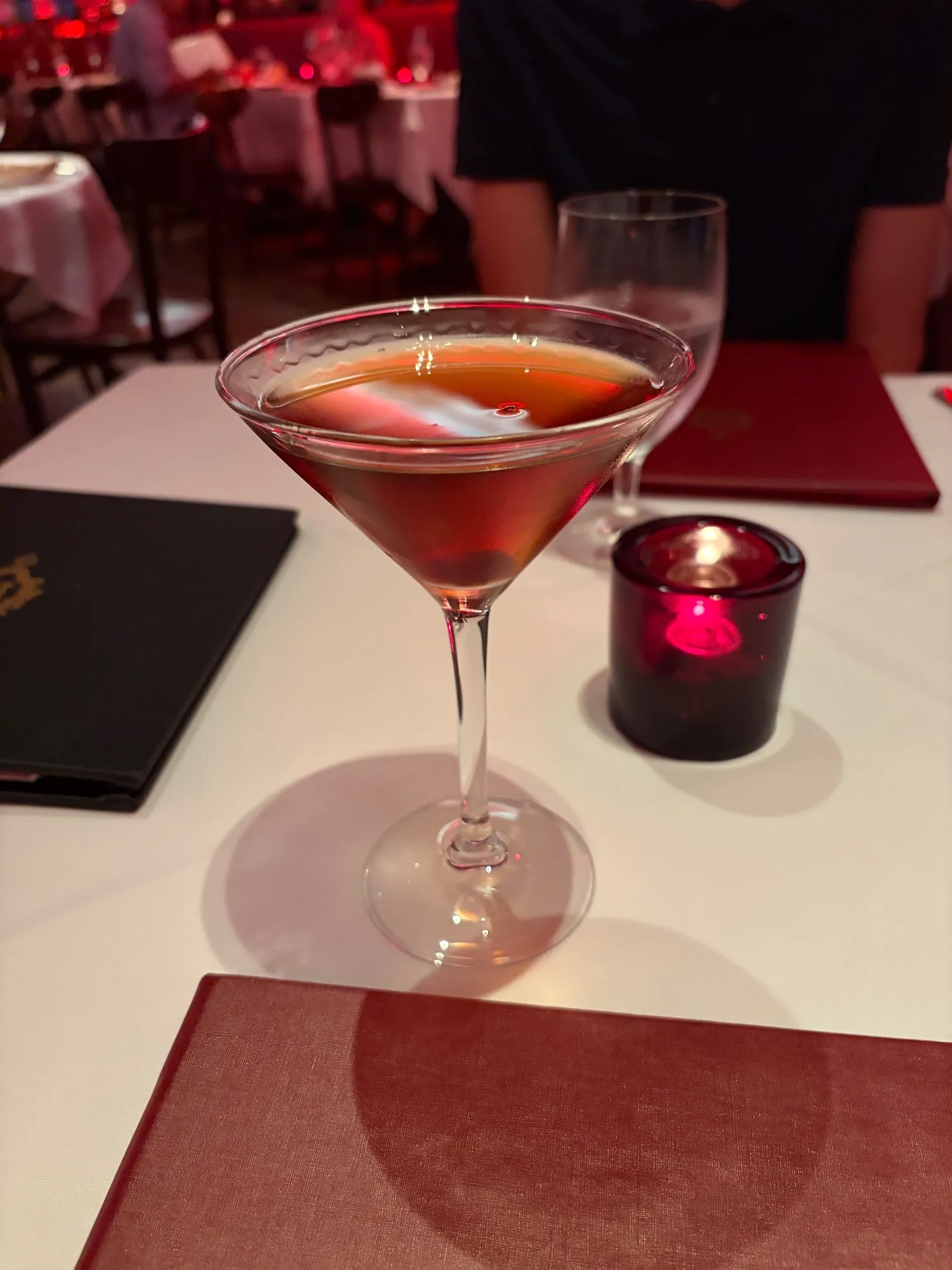 A brown cocktail served up in a stemmed glass on a white tablecloth.