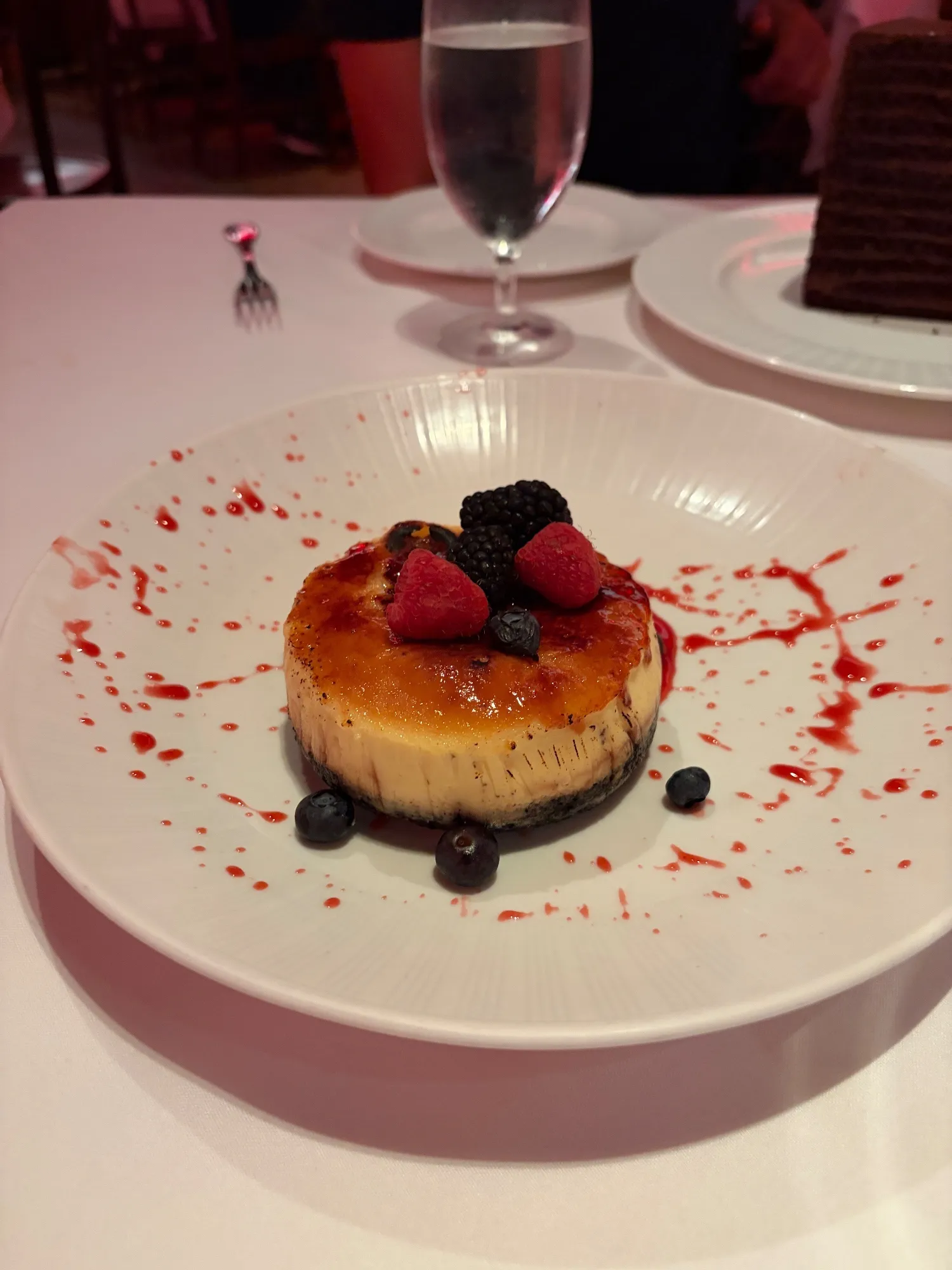 A round cheesecake with a caramelized top and dark crust, garnished with berries and red sauce.