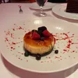 A round cheesecake with a caramelized top and dark crust, garnished with berries and red sauce.
